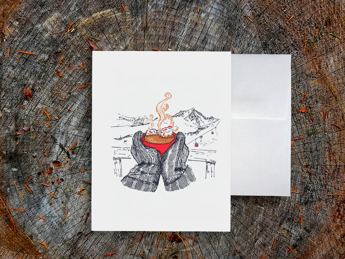A greeting card with an illustration of a red, steaming cup of hot chocolate being held by big mittens, with mountains in the background, on a wood surface next to a white envelope.