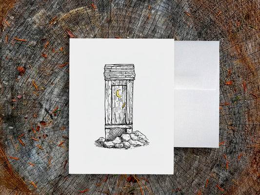 White card with a black ink drawing of an outhouse on a wooden surface.