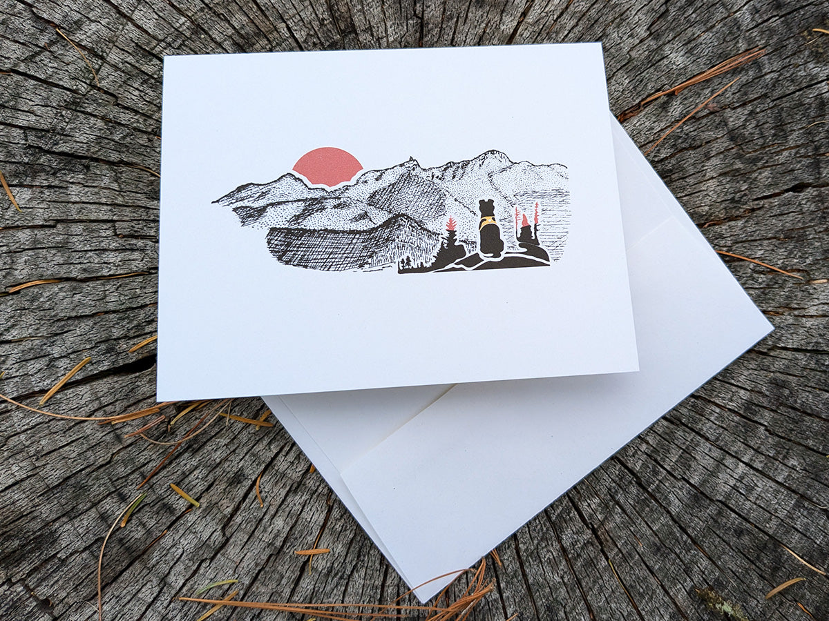 Greeting card with image of black dog looking at a red sunset behind mountains.