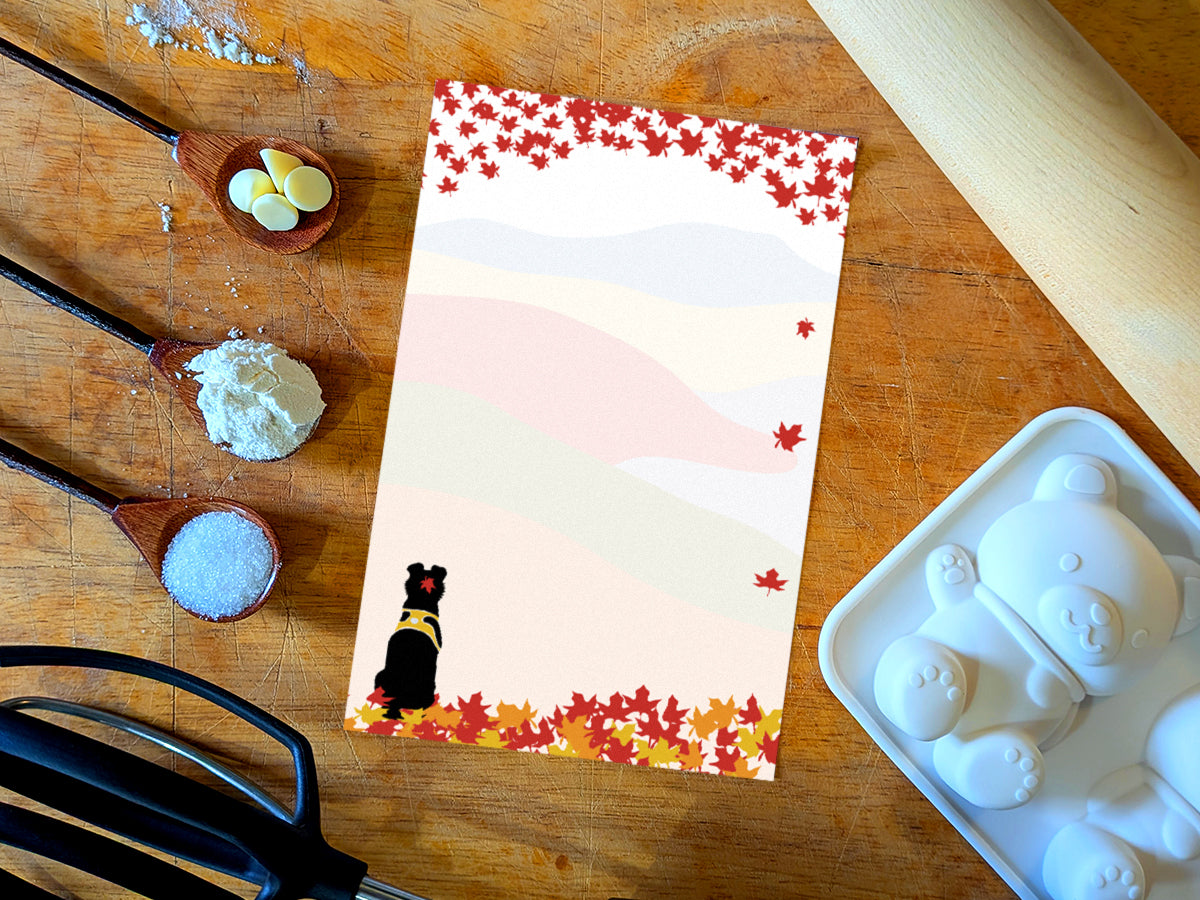 Notepad with an illustration of a black dog sitting among red, orange and yellow fall foliage with colorful rolling hills in the background, placed on a wooden surface with baking supplies.