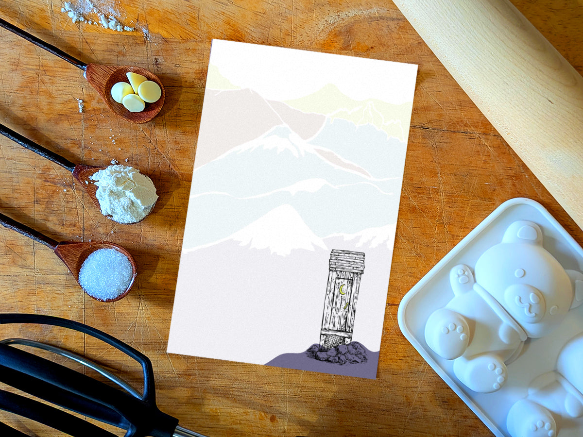 Notepad with an illustration of an outhouse in the mountains, placed on a wood surface with baking supplies.