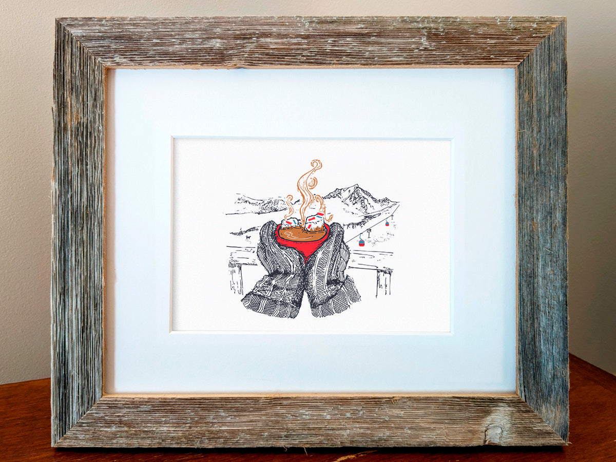 Framed art print illustration of thick winter mittens holding a red cup of hot chocolate, with mountains and chair lift in the background, displayed on a wooden shelf.