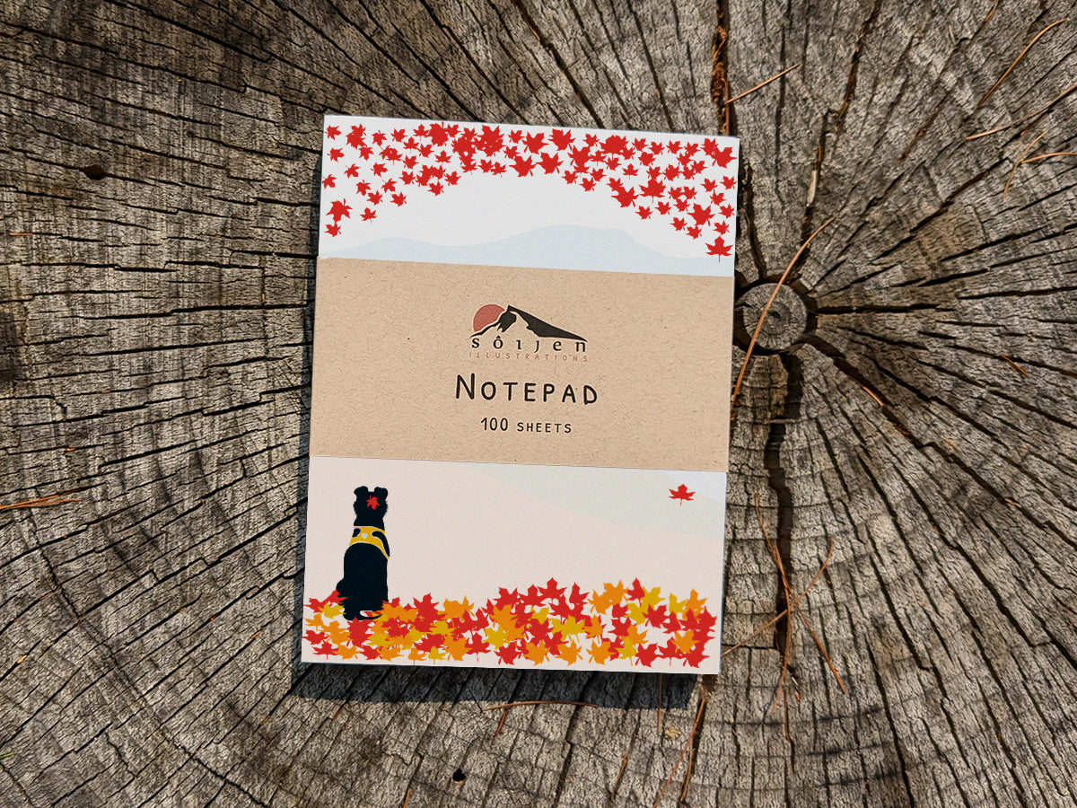 Notepad with a black dog among fall foliage on wooden background.