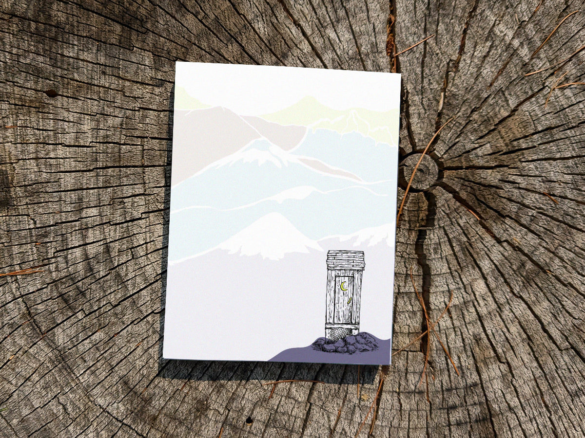 Notepad with an illustration of an outhouse in the mountains on a wood surface.
