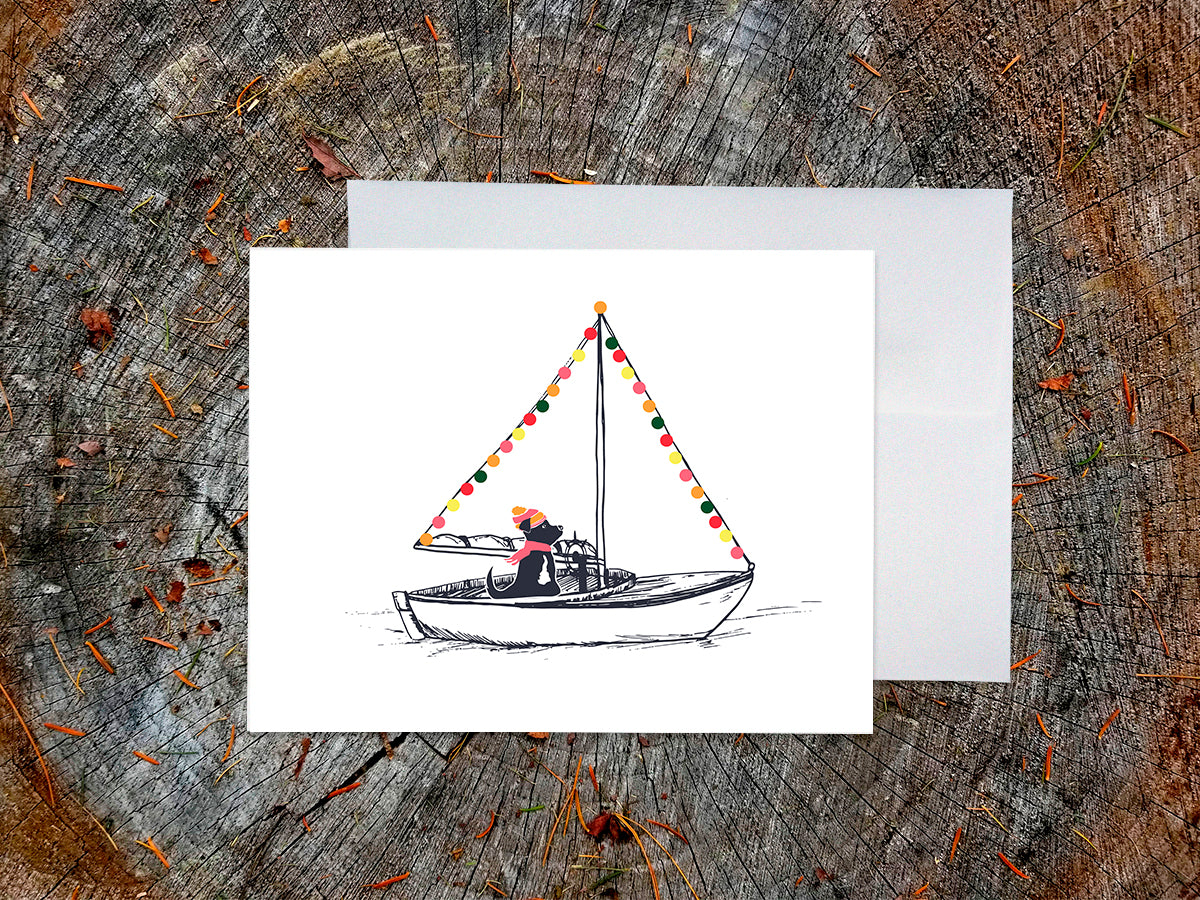 Greeting card with illustration of a black dog in a sailboat decorated with colorful lights, displayed on a wood surface with a white envelope.