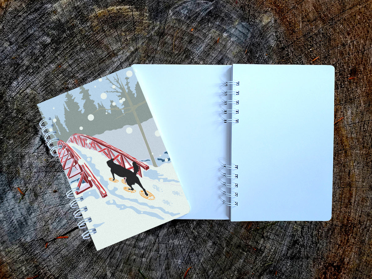 A spiral-bound notebook with an illustration of a dog snowshoeing over a bridge, shown on top of an open blank version of the notebook on a wood surface.