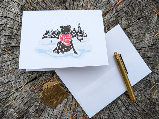 Greeting card with image of black dog with red scarf among a snowy winter landscape.