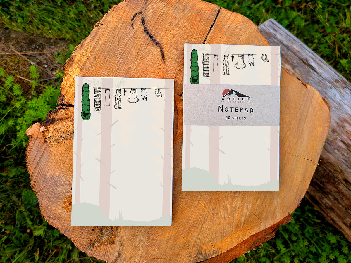 Two notepads with illustration of clothes and sleeping bag hanging on a line in between pine trees in a forest, placed on wooden surface.