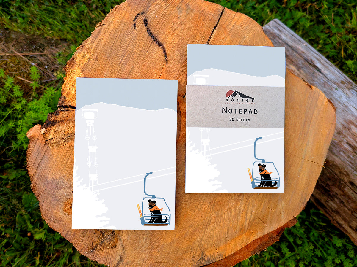 Two notepads with illustration of a dog with goggles and orange scarf riding a ski lift, placed on a wooden surface.
