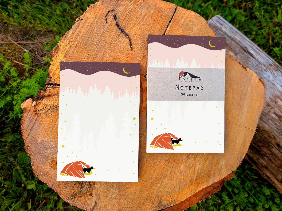 Two notepads with an illustration of a black dog in red tent camping under night sky with fireflies, placed on a wood surface.