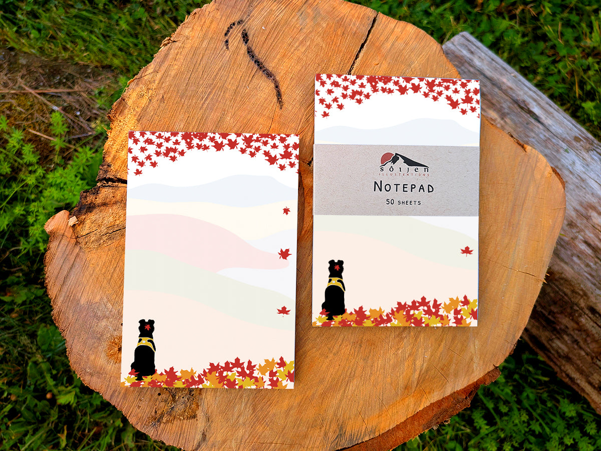 Two notepads with an illustration of a black dog sitting among red, orange and yellow fall foliage with colorful rolling hills in the background, placed on a wooden surface.