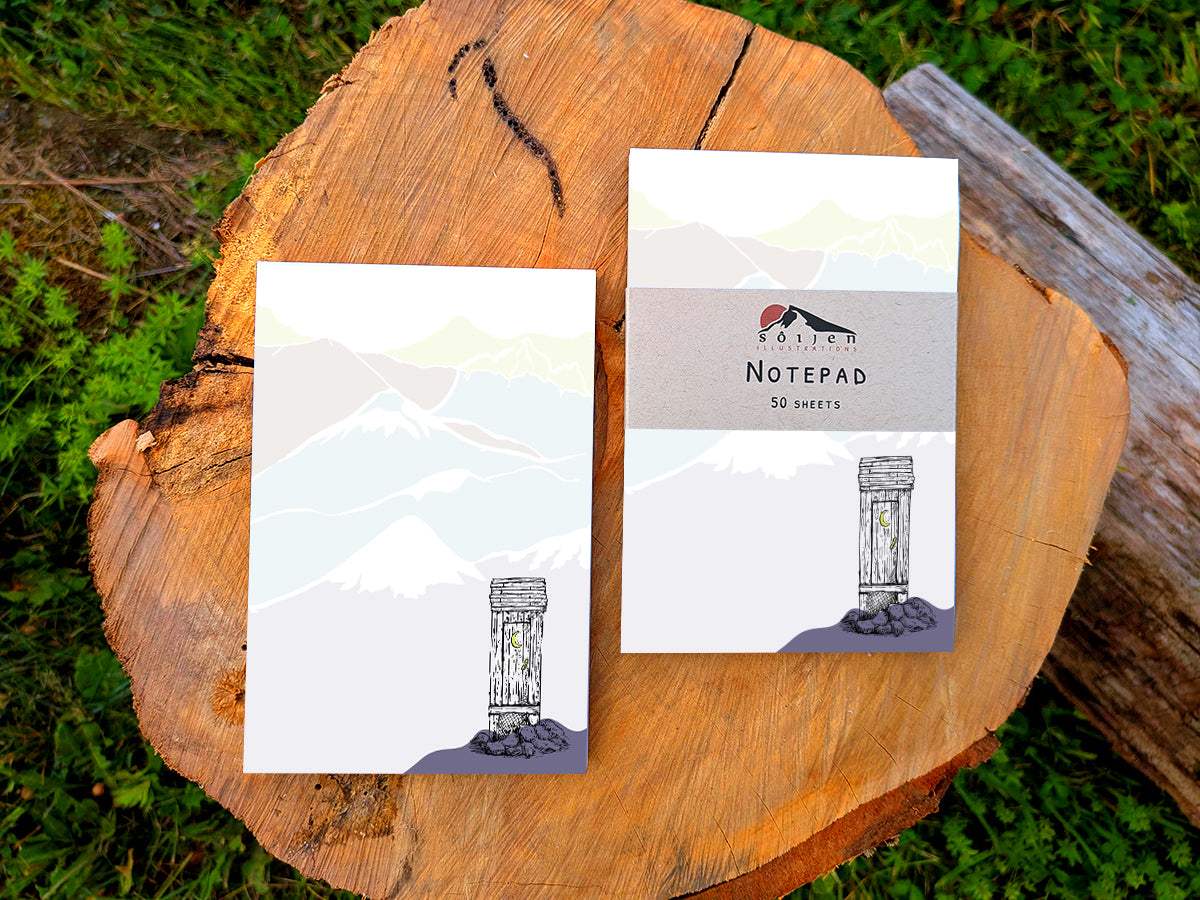 Two notepads with an illustration of an outhouse in the mountains, placed on a wood surface.