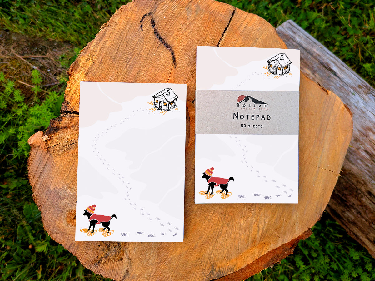 Two notepads with illustration of a black dog with winter hat, red jacket, and snowshoes, with a trail a footprints leading from a rustic cabin in the background, placed on a wooden surface.
