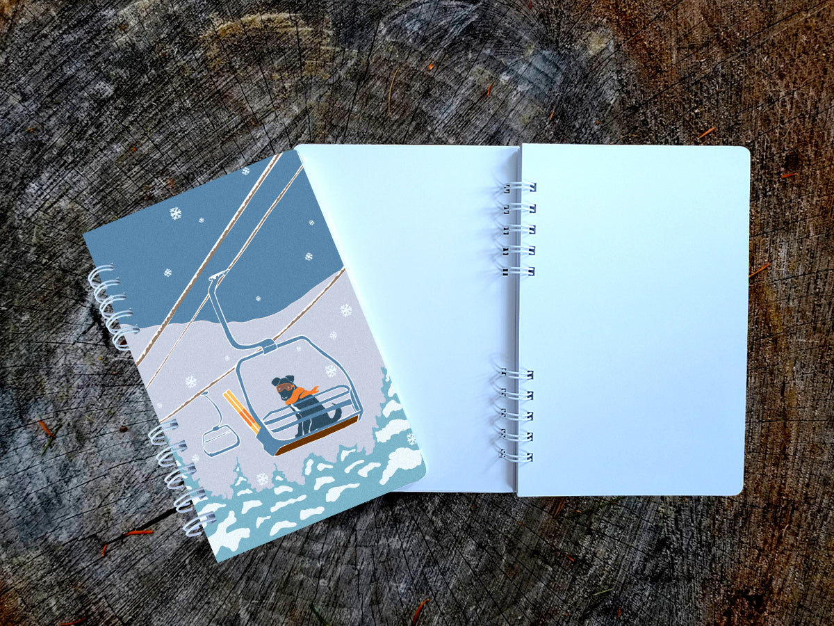 A spiral-bound notebook with an illustration of a dog riding a ski lift, shown on top of an open blank version of the notebook on a wood surface.