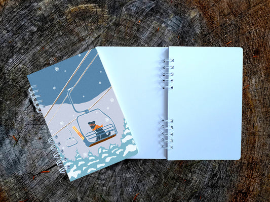 A spiral-bound notebook with an illustration of a dog riding a ski lift, shown on top of an open blank version of the notebook on a wood surface.