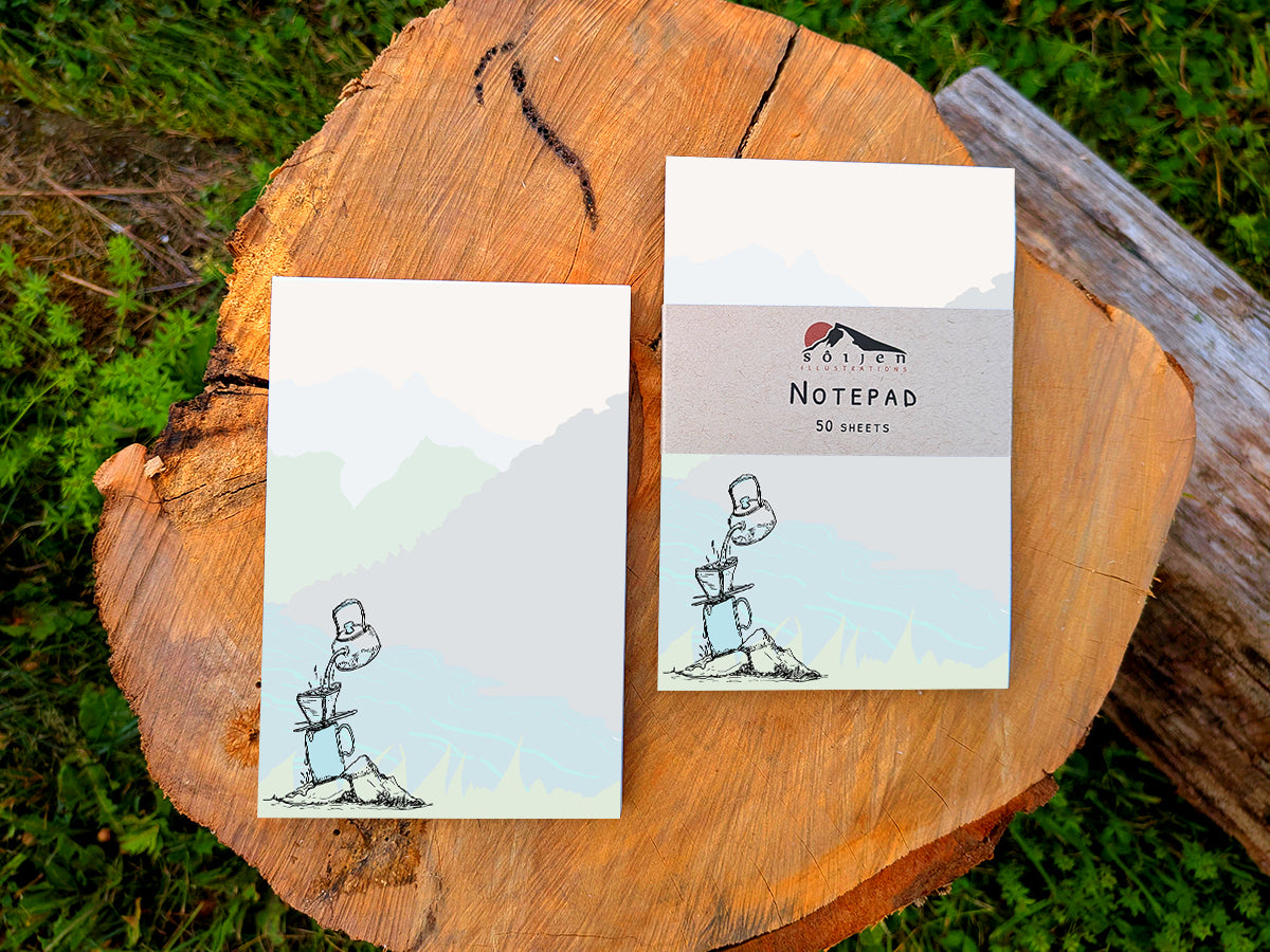 Two notepads with illustration of pour-over coffee on a riverbank, placed on a wood surface.