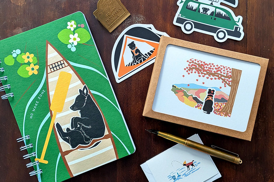 Notebook with dog and canoe design, cards, stickers, and pen on a wooden surface